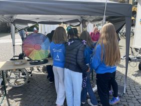 Ein Team von jungen Sportlerinnen stehen am Stand der LAG SH Sachsen beim Turnfest in Leipzig. Sie sind von hinten zu sehen und tragen blaue Sportjacken auf denen die Worte "Ingolstadt" und "Turnen" zu erkennen sind. 
