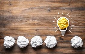 Gezeichnete Gl&uuml;hbirne mit Strahlen und daneben in einer Linie zerkn&uuml;llte Papierkugeln auf einem Holzhintergrund