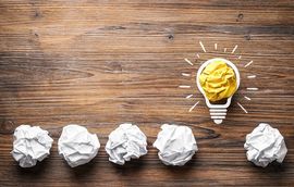 Gezeichnete Gl&uuml;hbirne mit Strahlen und daneben in einer Linie zerkn&uuml;llte Papierkugeln auf einem Holzhintergrund