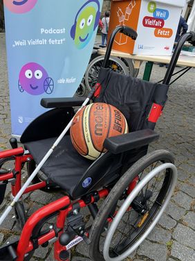 Zu sehen ist ein Rollstuhl am Stand der LAG SH Sachsen beim Turnfest in Leipzig. Auf dem Rollstuhl liegen ein Volleyball und ein Blindenlangstock. Im Hintergrund sieht man die Aufsteller der Projekte Challenge Inklusion und Weil Vielfalt fetzt.