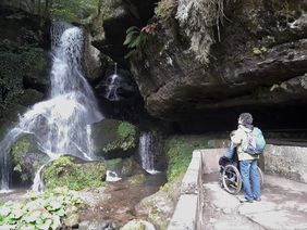 Rollstuhlnutzer mit Begleitperson von hinten stehen vor einem Wasserfall in Bad Schandau