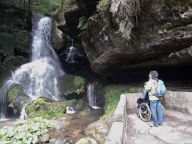 Rollstuhlnutzer mit Begleitperson von hinten stehen vor einem Wasserfall in Bad Schandau