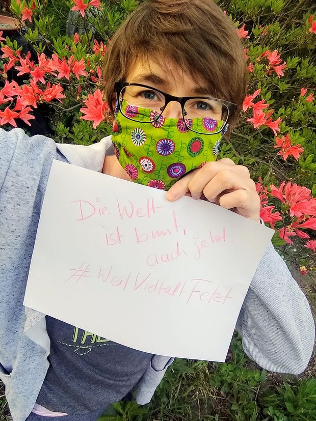 Susanne Rößner hält ein Zettel auf dem steht: Die Welt ist bunt, auch jetzt! #WeilVielfaltFetzt. Sie trägt eine bunte Atamschutzmaske und steht vor einem blühenden Busch
