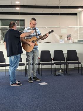 Stephan Walther spielt auf seiner Gitarre und singt. Er trägt einen Irokesenschnitt. Ein Blatt mit Noten und Text liegt vor ihm auf dem Boden. Ein anderer Mann hält ihm das Mikrofon vor den Mund.