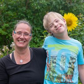 Mutter mit Kind fr&ouml;hlich lachend, Kind umarmt Mutter seitlich, sie stehen hinter einem Sonnenblumenfeld