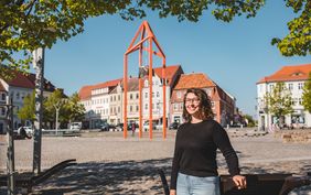 Frau mit schwarzem-lockigem Haar und Brille im schwarzen Shirt steht auf dem Marktplatz von Bischofswerda, Frau l&auml;chelt in die Kamera