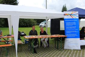 Zwei Mitarbeiter des Inklusionsnetzwerk Sachsen, Falk Friedrich (links) und Angela Werner (rechts) sitzen an einem Tisch am Stand des INWS. Auf dem Tisch liegen Bauteile eines Mobilitätsbaukastens für Rollstuhlfahrer. Die beiden sitzen unter einem Zeltdach an dem ein Roll-Up des INWS befestigt ist.