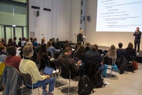 Blick über die Zuschauer auf das Podium des Netzwerktreffens. Frau Sappinen steht am Podium und referiert. Im Hintergrund ist ihre Präsentation auf einer großen Leinwand zu sehen.