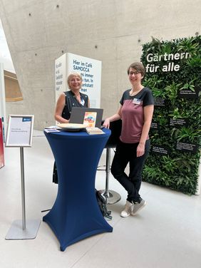 Katja und Susanne Rößner stehen an einem Stehtisch und lächeln in die Kamera. Links daneben steht ein Schild "Erkundung Wanderausstellung". Hinter ihnen befinden sich zwei Stelen der Wanderausstellung "Weil Vielfalt fetzt". Eine Stele sieht so aus als würde sie aus Pflanzen bestehen und trägt die Aufschrift "Gärtnern für alle". Auf der anderen kann man verschiedene Texte erahnen.