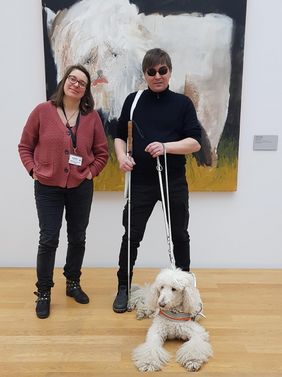 Katrina Blach und Sebastian Schulze stehen nebeneinander vor einem großen Gemälde eines weißen Büffels des Neo-Rauch-Schülers Julius Hofmann im Mdbk. Katrina Blach trägt eine rote Strickjacke und eine schwarze Jeans. Sie hat die Hände entspannt in den Hosentaschen und lächelt in die Kamera. Sie trägt ihre braunen Haare schulterlang und eine Brille. Sebastian Schulze ist ganz in schwarz und trägt eine Sonnenbrille. In den Händen hält er einen Langstock und die Leine seines Hundes. Vor ihnen liegt der weiße Blindenhund von Sebastian Schulze.