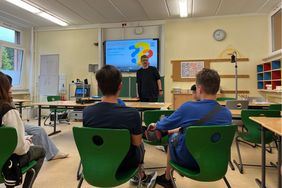 Ein Mann referiert vor Sch&uuml;ler*innen in einem Klassenraum. Er hat einen Bart, ist Ende 30, hat eine Brille und tr&auml;gt schwarze Kleidung. Drei Sch&uuml;ler, ein M&auml;dchen und 2 Jungen, sind von hinten zu sehen w&auml;hrend sie an ihren Schultischen sitzen. An der Tafel ist eine Powerpoint Pr&auml;sentation zum Thema Inklusion zu sehen. 