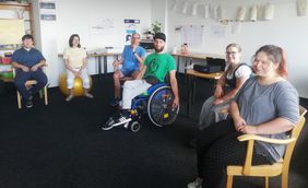 Menschen sitzen im Halbkreis auf einen Stuhl und blicken in die Kamera, sitzende Perspektiv