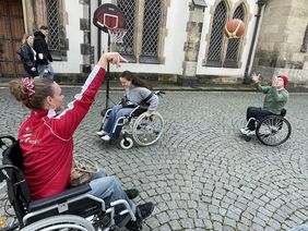 Zwei junge Damen und ein junger Mann sitzen in Rollst&uuml;hlen und spielen Rolstuhlbasketball. Die Dame im Vordergrund passt den Ball zu dem jungen Mann. Der Ball befindet sich in der Luft zwischen den beiden Spielenden. Im Hintergrund f&auml;hrt die zweite Dame mit dem Rollstuhl umher. 