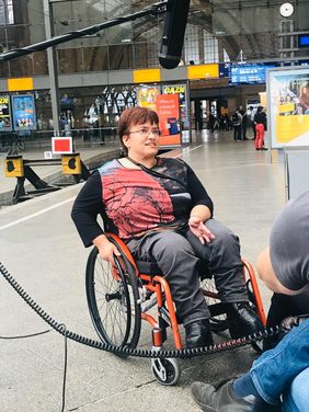 Frau mit kurzen Haaren und Brille im Rollstuhl am Bahnsteig im Interview