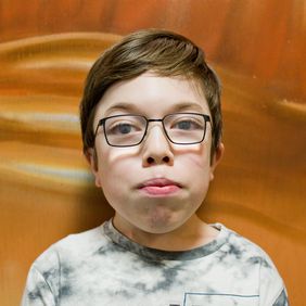 Lukas blickt mit ernster Mine in die Kamera. Er hat eine Brille auf, die seine Augen etwas vergrößern. Er hat kurze braune Haare. 