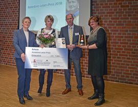 Vier Menschen, darunter die zwei Gewinner mit gro&szlig;em Scheck und Blumenstrau&szlig; in der Hand