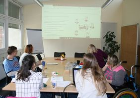 Teilnehmer sitzen am Tisch und schauen zur Leinwand