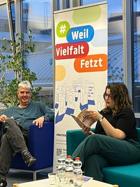 Birger H&ouml;hn und Marsha Richarz sitzen auf Sesseln neben dem Aufsteller mit der Aufschrift "Weil Vielfalt fetzt". Marsha Richarz liest konzentriert in dem Buch, Birger H&ouml;hn l&auml;chelt jemanden au&szlig;erhalb des Bildes an.