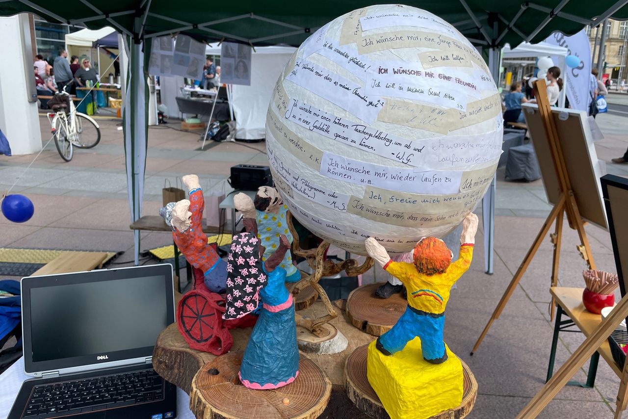 An einem Stand steht eine Weltkugel mit vielen Wünschen darauf. Sie wird von Figuren verschiedener ethnischer Abstammung und mit und ohne Behinderung gehalten.