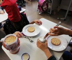 Tisch mit Geschirr, Brot und Frischkäse. Zwei Kinder versuchen mit verbundenen Augen das Brot zu schmieren.