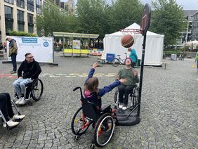 Ein junges M&auml;dchen sitzt im Rollstuhl und wirft einen Korb beim Rollstuhl-Basketball. Zwei junge M&auml;nner Anfang Zwanzig sitzen im Rollstuhl daneben. Alle sehen sehr erfreut aus und haben Spa&szlig;.