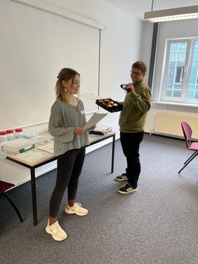 Lara Hanske steht vor den Teilnehmer*innen und liest etwas von einem Zettel ab. Sie trägt ihre halblangen blonden Haare in einem Zopf und hat eine schwarze Hose und ein graues Oberteil an. Stephan Zapke hält verschiedene Simulationsbrillen in der Hand. Er ist sichtbar sehbehindert und trägt eine schwarze Hose und einen grünen Pullover. Er hat kurze dunkelblonde Haare.