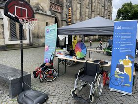 Der Stand der LAG SH Sachsen beim Turnfest in Leipzig. Zu Sehen ist ein Pavillon mit einer Biertischgarnitur auf der ein Gl&uuml;cksrad steht und verschiedene Flyer ausliegen. Vor dem Stand stehen zwei Rollst&uuml;hle und ein Basketballkorb. Im Hintergrund sieht man die Thomaskirche in Leipzig.