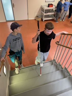 Zwei Jungen einer Grundschulklasse gehen eine Treppe hinauf. Einer der Jungen tr&auml;gt eine Augenmaske und hat einen Blindenlangstock in der Hand. Er h&auml;lt sich am Gel&auml;nder fest, w&auml;hrend er versucht blind die Treppe hinaufzusteigen. Der zweite sehende Junge steht neben ihm und passt auf den anderen jungen mit dem Blindenlangstock auf.