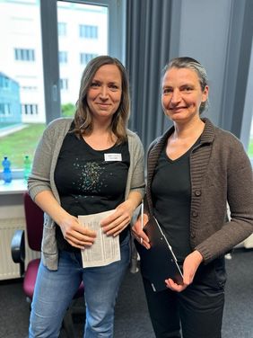 Kerstin Helm und Heike Engelien lächeln in die Kamera. Kerstin Helm hat halblange braune Haare, trägt eine graue Strickjacke, ein schwarzes Tshirt und Jeans. Heike Engelien trägt ihre dunklen Haare im Zopf und hat eine schwarze Hose, ein schwarzes Tshirt und eine braune Strickjacke an. Beide halten Zettel mit Quizaufgaben in der Hand.