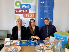 Das Team der LAG SH Sachsen sitzt am Stand der LAG SH Sachsen auf dem Bildungskongress in Görlitz. Katja Rößner (links), Kerstin Helm (mitte) und Stephan Zapke (rechts). Auf dem Tisch vor dem Team liegen verschiedene Materialen aus den Projekten der LAG SH Sachsen aus.