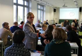 Annegret Haage h&auml;lt ein Mikrofon in Richtung eines Teilnehmers und steht mitten zwischen den Tischen mit Teilnehmenden