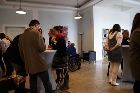 Fraauen und M&auml;nner stehen um Stehtischen und sprechen miteinander