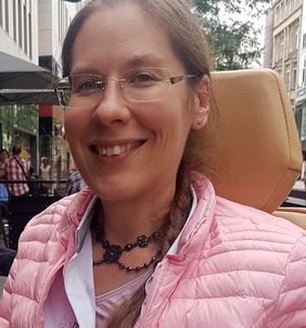 l&auml;chelnde Frau mit Brille, gebundenen Haaren sitzend im Rollstuhl mit Schmuck um den Hals