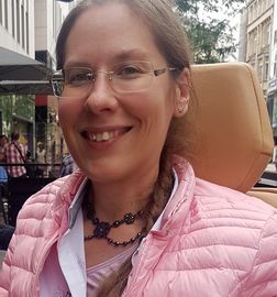 l&auml;chelnde Frau mit Brille, gebundenen Haaren sitzend im Rollstuhl mit Schmuck um den Hals