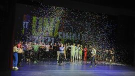 Viele junge Menschen stehen auf einer Theaterbühne. Die Scheinwerfer leuchten hell und hinter der Gruppe steht sehr groß Unart an der Wand. Einige tragen Kostüme, andere haben sich einfach schick gemacht. Einzelheiten lassen sich schlecht erkennen. Die Jugendlichen stehen in einem bunten Konfettiregen. Er regnet von oben auf Bühne herab. Alle freuen sich. Viele von ihnen klatschen in die Hände. Es ist der Abschluss des Unart-Finale 2022 im Kleinen Haus. 