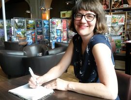 Frau mit Brille, halb langen Haaren am Tisch sitzend und Stift und Notitzbuch dabei