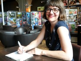 Frau mit Brille, halb langen Haaren am Tisch sitzend und Stift und Notitzbuch dabei