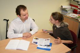 Beratungsszene - Mann und Frau im Gespr&auml;ch, davon einer mit Gehhilfe sitzend am Tisch