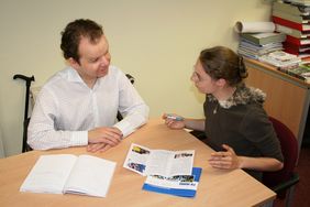 Beratungsszene - Mann und Frau im Gespr&auml;ch, davon einer mit Gehhilfe sitzend am Tisch